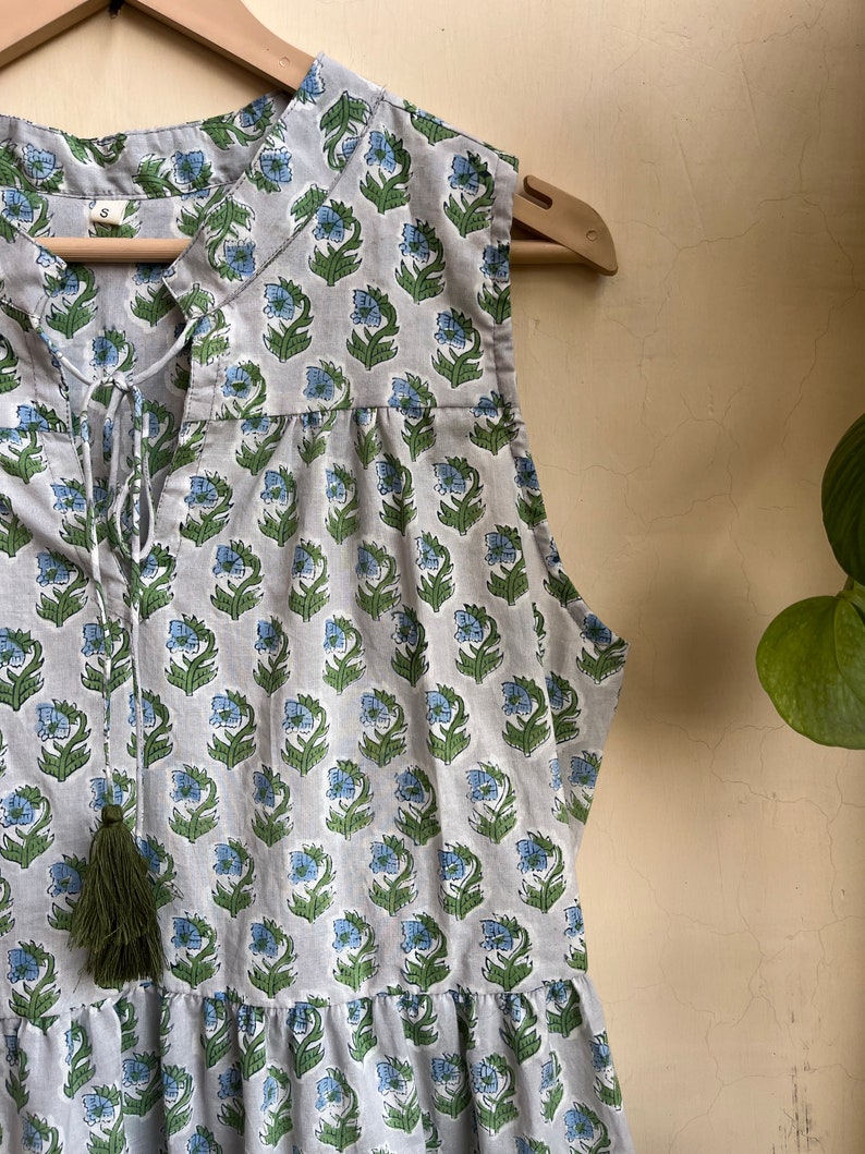 Op de afbeelding: Een grijs, mouwloos jurkje met een bloemenprint in blauwe en groene tinten. Het jurkje heeft een strik aan de halslijn en een kwast.