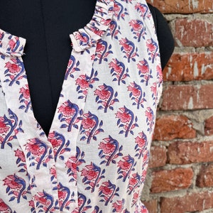 May include: White sleeveless dress with a floral pattern in pink and blue. The dress features a ruffled collar and a V-neckline. The fabric appears to be lightweight cotton, ideal for warm weather.