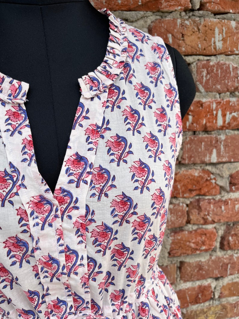 May include: White sleeveless dress with a floral pattern in pink and blue. The dress features a ruffled collar and a V-neckline. The fabric appears to be lightweight cotton, ideal for warm weather.