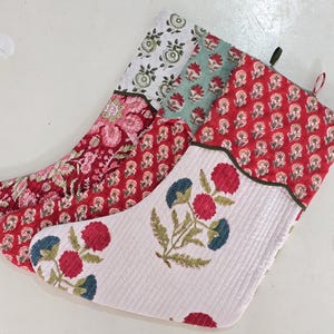 Puede incluir: Tres calcetines navideños acolchados con estampados florales. Uno es blanco con flores rojas y azules, otro es rojo con pequeñas flores blancas, y el tercero es una mezcla de estampados florales rojos y rosas. Cada uno tiene un ribete verde oscuro.
