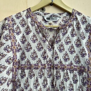 May include: A white cotton dress with a purple and brown floral print. The dress has a V-neckline and a tie at the neck. The dress has a pleated bodice and a gathered skirt. The dress has a label that says "Boh Creation Clothing" and a size "S".
