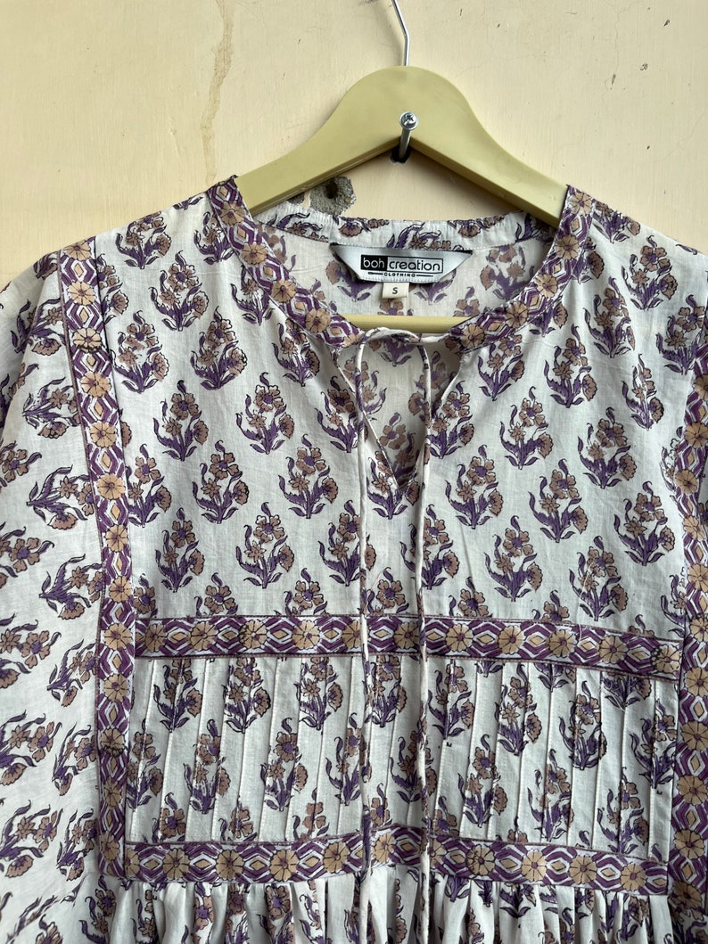 May include: A white cotton dress with a purple and brown floral print. The dress has a V-neckline and a tie at the neck. The dress has a pleated bodice and a gathered skirt. The dress has a label that says "Boh Creation Clothing" and a size "S".