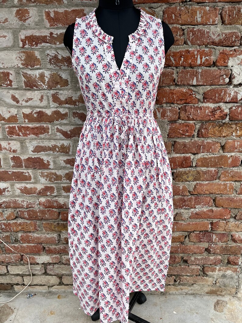May include: White sleeveless dress with a floral pattern in red and blue. The dress has a V-neckline, a drawstring waist, and a knee-length skirt. The dress is displayed on a black mannequin.