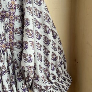 May include: A white cotton dress with a floral print in shades of purple and brown. The dress has a gathered bodice and long sleeves with elastic cuffs.