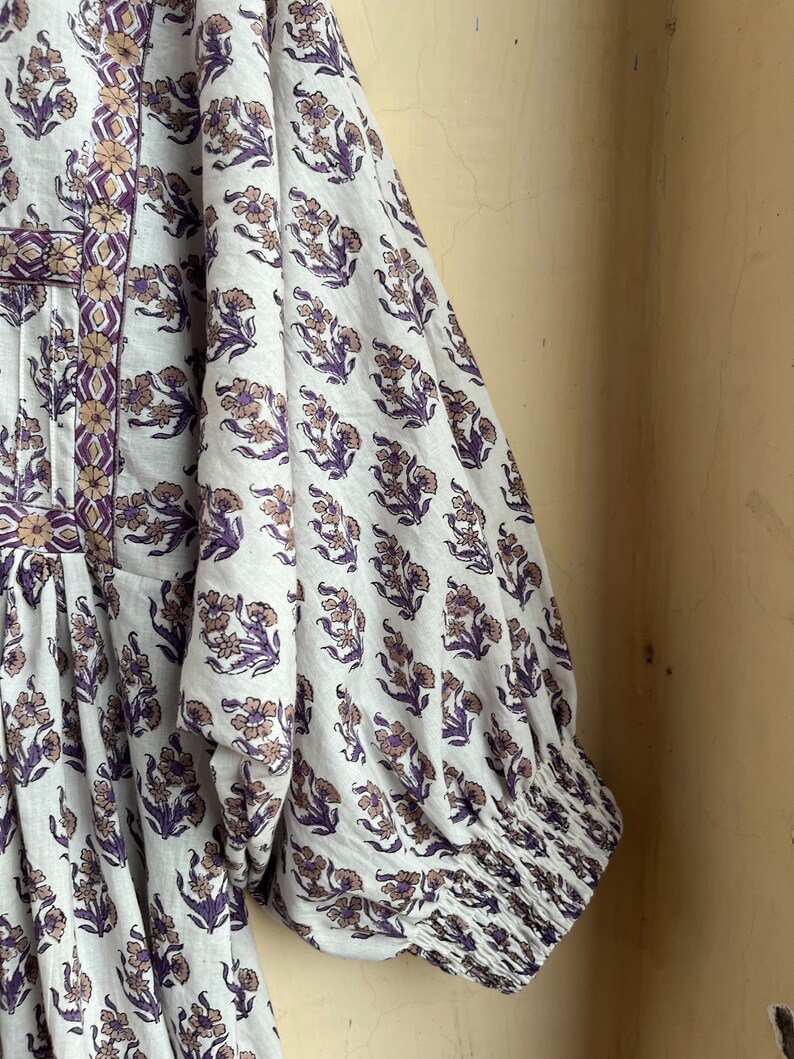 May include: A white cotton dress with a floral print in shades of purple and brown. The dress has a gathered bodice and long sleeves with elastic cuffs.