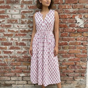 May include: A sleeveless white dress with a pink floral pattern. The dress has a V-neck and a gathered waist. The model is wearing brown sandals. The background is a red brick wall.