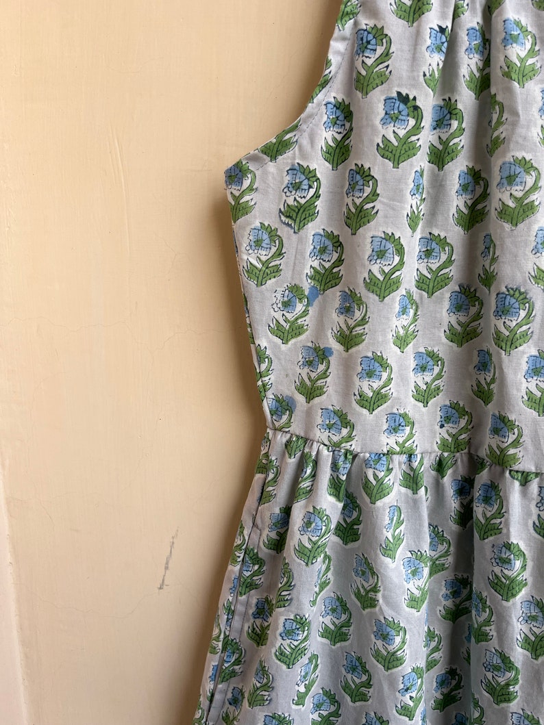 Op de afbeelding: Een mouwloze jurk met een lichtblauwe en groene bloemenprint. De jurk heeft een geplooide rok en een halternek.