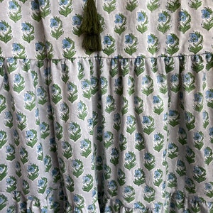 Op de afbeelding: Een witte katoenen jurk met een blauwe en groene bloemenprint. De jurk heeft een gelaagde rok en een groene kwast aan de halslijn.