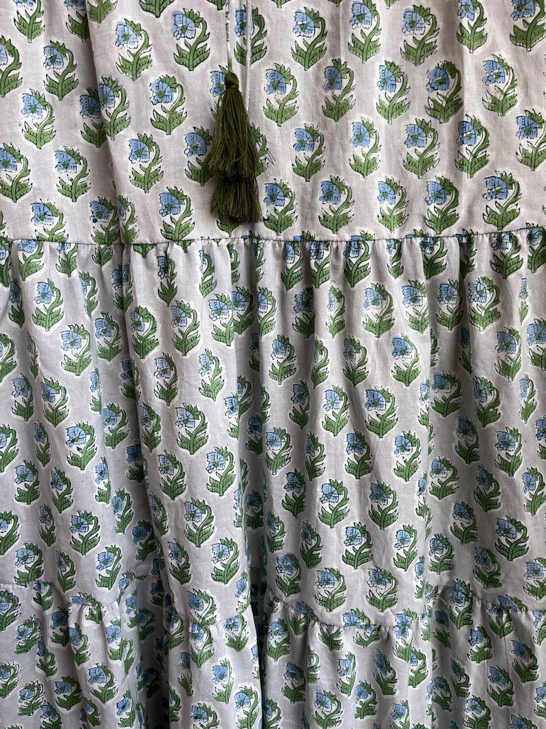 Op de afbeelding: Een witte katoenen jurk met een blauwe en groene bloemenprint. De jurk heeft een gelaagde rok en een groene kwast aan de halslijn.