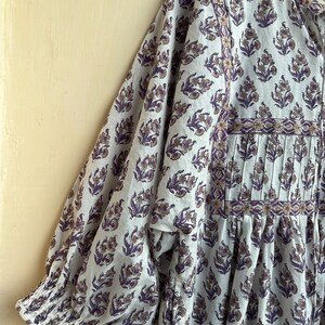 May include: A white cotton dress with a purple floral print and a gathered skirt. The dress has a round neckline and 3/4 sleeves with elastic cuffs.