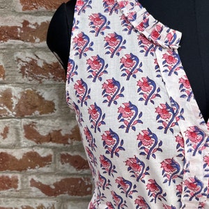 May include: Sleeveless white dress with a floral pattern. The dress features a V-neckline and is adorned with a repeating design of pink flowers and blue stems. The fabric appears to be lightweight, suitable for warm weather.
