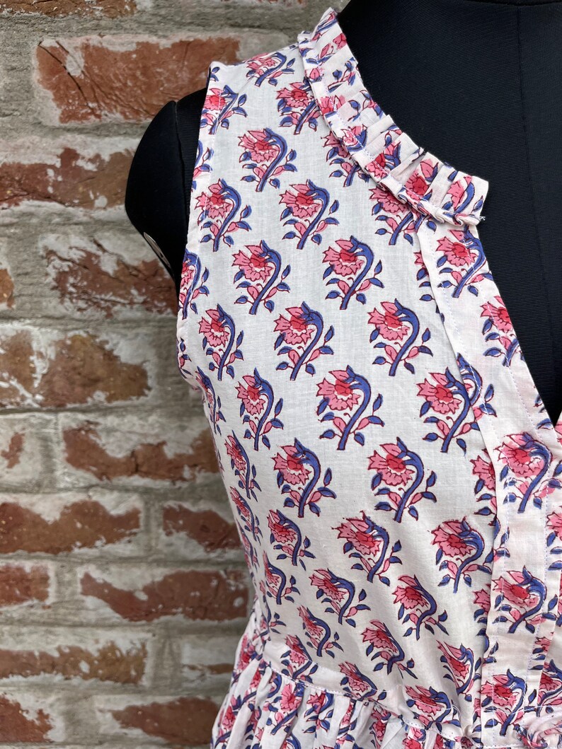 May include: Sleeveless white dress with a floral pattern. The dress features a V-neckline and is adorned with a repeating design of pink flowers and blue stems. The fabric appears to be lightweight, suitable for warm weather.