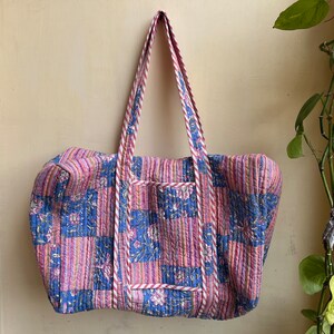 May include: A large, quilted tote bag with a patchwork design. The bag features a combination of pink, blue, and white floral patterns. The handles and trim are striped in pink and white. The bag is hanging against a light-colored wall.