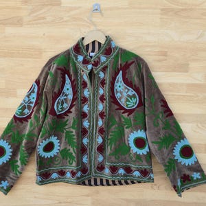 Può includere: Giacca in velluto marrone con ricami intricati. La giacca presenta motivi floreali e paisley verdi, bordeaux e azzurri. È appesa a una gruccia di legno su uno sfondo di legno.