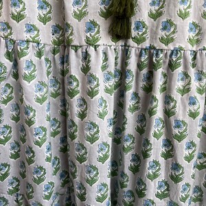 Op de afbeelding: Een witte katoenen jurk met een blauwe en groene bloemenprint. De jurk heeft een gelaagde rok en een kwastdetail aan de halslijn.