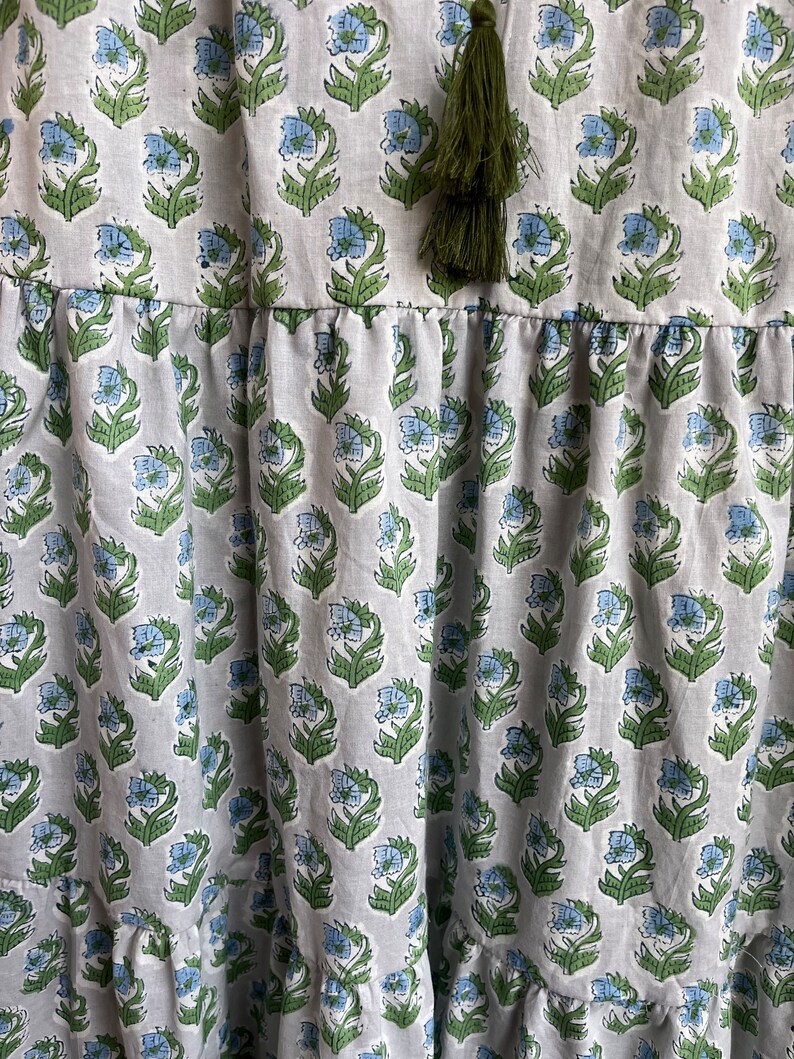 Op de afbeelding: Een witte katoenen jurk met een blauwe en groene bloemenprint. De jurk heeft een gelaagde rok en een kwastdetail aan de halslijn.