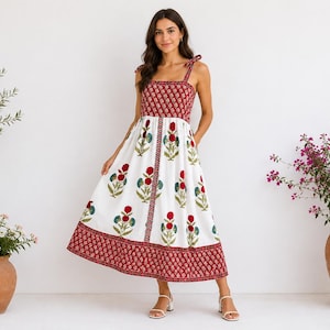 Op de afbeelding: Witte midi-jurk met een rode bloemenprint en een rood patroon op het lijfje en de zoom. De jurk heeft strikbandjes en wordt gedragen met witte sandalen. De achtergrond is een witte muur met potplanten aan weerszijden.