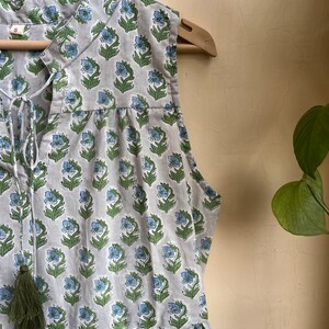 Op de afbeelding: Een mouwloze grijze jurk met een bloemenprint in tinten blauw en groen. De jurk heeft een V-hals met een strik sluiting en een kwast detail aan de hals.