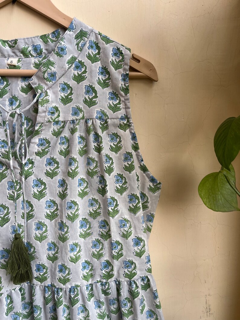 Op de afbeelding: Een mouwloze grijze jurk met een bloemenprint in tinten blauw en groen. De jurk heeft een V-hals met een strik sluiting en een kwast detail aan de hals.