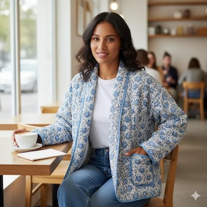 May include: A blue and white floral quilted jacket with light blue trim. The jacket is worn over a white t-shirt and blue jeans. The jacket has two pockets. The setting is a cafe.
