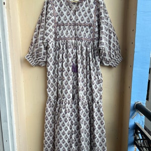 May include: A white cotton dress with a purple floral print. The dress has a gathered waist, a round neckline, and 3/4 sleeves. The dress is embroidered with purple and orange thread.