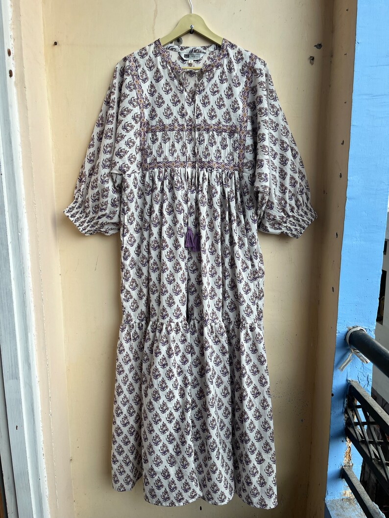 May include: A white cotton dress with a purple floral print. The dress has a gathered waist, a round neckline, and 3/4 sleeves. The dress is embroidered with purple and orange thread.