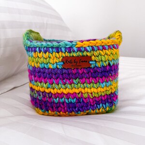 May include: A colourful crocheted basket with a brown leather tag that says "Knots by Emma". The basket is made with a variety of bright colours, including yellow, green, purple, and blue.