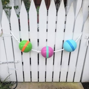 May include: A white picket fence with a string of three colorful Easter eggs hanging from it. The eggs are green, pink, and blue with white stripes.