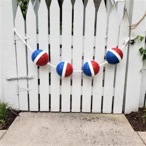 May include: A red, white, and blue nautical rope garland with four buoys hanging from it. The garland is strung across a white picket fence.