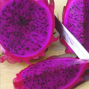 Puede incluir: Primer plano de un fruto del drag&oacute;n morado cortado por la mitad, revelando las semillas negras en el interior. La fruta se est&aacute; cortando con un cuchillo plateado sobre una superficie de madera.