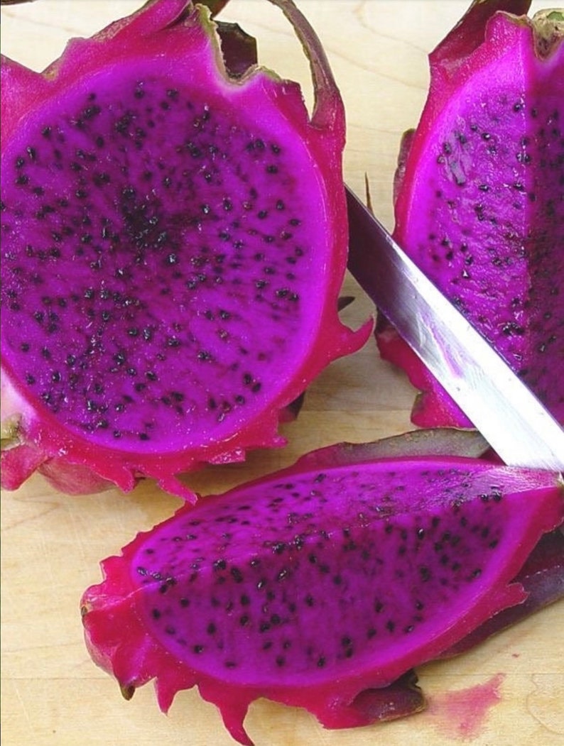 Puede incluir: Primer plano de un fruto del drag&oacute;n morado cortado por la mitad, revelando las semillas negras en el interior. La fruta se est&aacute; cortando con un cuchillo plateado sobre una superficie de madera.