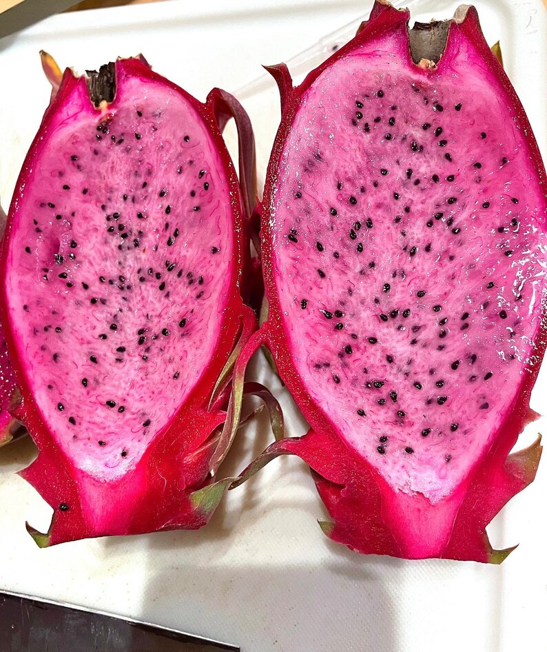 Puede incluir: Dos mitades de fruta de drag&oacute;n rosa con semillas negras. La fruta est&aacute; cortada por la mitad y la pulpa es visible.