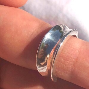 Puede incluir: Un anillo de plata con una banda ancha y curva. La banda es lisa y pulida, y tiene un borde ligeramente redondeado.