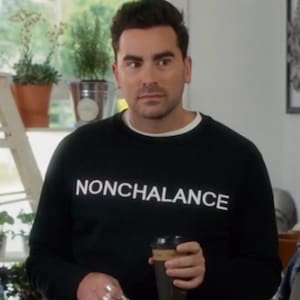 May include: A man wearing a black sweatshirt with the word "NONCHALANCE" printed in white on the front. He is holding a brown paper cup of coffee.