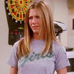 May include: A woman with long blonde hair is wearing a grey t-shirt with the word "girls" written in green on the front.