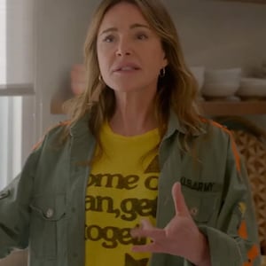 May include: A woman wearing a green jacket with orange stripes and a yellow t-shirt with the text "come on an, get toget" in brown lettering. The t-shirt also has a brown star and a small black patch that says "US ARMY".