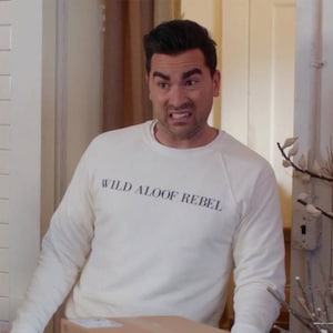 May include: A man wearing a white sweatshirt with the text "WILD ALOOF REBEL" printed on the front. He is looking at the camera with a surprised expression.