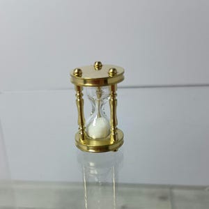 Puede incluir: Un pequeño reloj de arena decorativo con un marco de metal dorado. El reloj de arena tiene un centro de vidrio transparente lleno de arena blanca. La parte superior e inferior son circulares, y el marco tiene cuatro soportes verticales.