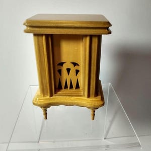 Puede incluir: Un modelo de chimenea de madera clara. De forma rectangular, presenta columnas verticales y cortes decorativos. Se apoya sobre cuatro pequeñas patas torneadas, sugiriendo un objeto decorativo.