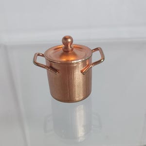 以下が含まれることがあります： 蓋と2つの小さな取っ手が付いた、銅色のミニチュア鍋。 鍋は円筒形で、垂直線と蓋に小さなノブが付いています。 ドールハウスやミニチュアコレクションに適した装飾品です。