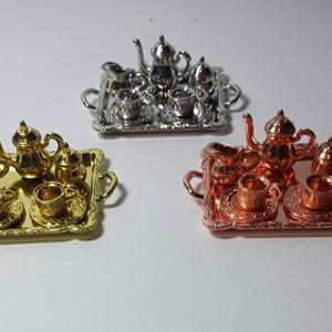 May include: Three miniature tea sets in silver, gold, and copper tones. Each set includes a rectangular tray with handles, a teapot, a creamer, a sugar bowl, and two teacups on saucers. The metal pieces feature embossed detailing on the cups and trays.