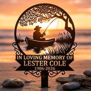 Op de afbeelding: Zwart metalen gedenkteken met een silhouet van een persoon die vanuit een boot onder een boom vist. Het bord leest "IN LOVING MEMORY OF LESTER COLE 1986-2026" tegen een zonsondergang achtergrond. Het bord staat op een zandstrand.