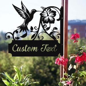 May include: Black metal sign with a hummingbird and hibiscus flower design. The sign has the text "Custom Text" in a decorative font. Suitable for outdoor use, the sign is mounted on a wooden post.