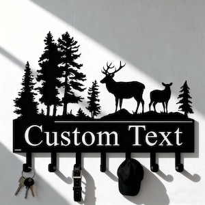 Puede incluir: Llavero de metal negro con un diseño de escena de bosque, con ciervos y árboles. Tiene ganchos para llaves, cinturones y sombreros, y el texto "Custom Text". Un accesorio decorativo y funcional.