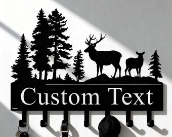 Pode incluir: Porta-chaves de metal preto com um design de cena de floresta, com veados e árvores. Possui ganchos para chaves, cintos e chapéus, e o texto "Custom Text". Um acessório doméstico decorativo e funcional.