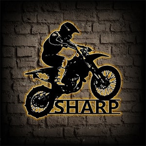 Op de afbeelding: Zwarte silhouet van een persoon die een crossmotor rijdt, springend over het woord "SHARP" in geel.