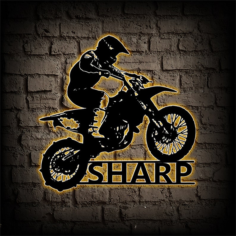 Op de afbeelding: Zwarte silhouet van een persoon die een crossmotor rijdt, springend over het woord "SHARP" in geel.