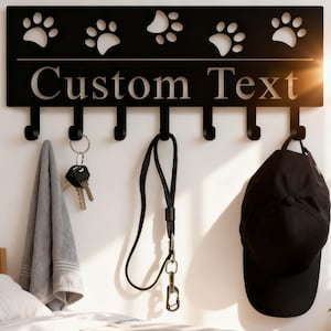 Pode incluir: Organizador de parede em metal preto com recortes de pegadas e ganchos. A placa diz "Custom Text" e contém uma toalha, chaves, uma trela e um boné preto. Um item decorativo e funcional para donos de animais.
