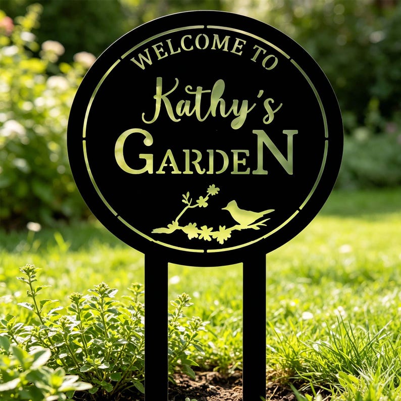 Puede incluir: Letrero de jard&iacute;n de metal negro con el texto "WELCOME TO Kathy's GARDEN" en un dise&ntilde;o circular. El letrero presenta una ilustraci&oacute;n de un p&aacute;jaro y una rama floral. El letrero est&aacute; colocado en un jard&iacute;n con hierba y plantas verdes.