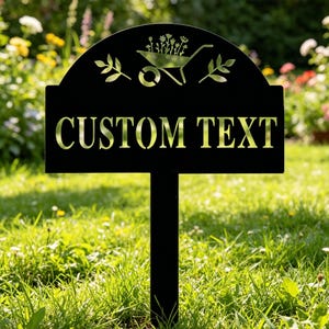Puede incluir: Letrero de jard&iacute;n de metal negro con las palabras "CUSTOM TEXT" recortadas. La parte superior del letrero presenta una carretilla y un dise&ntilde;o floral. El letrero est&aacute; clavado en un c&eacute;sped con un fondo borroso de flores.